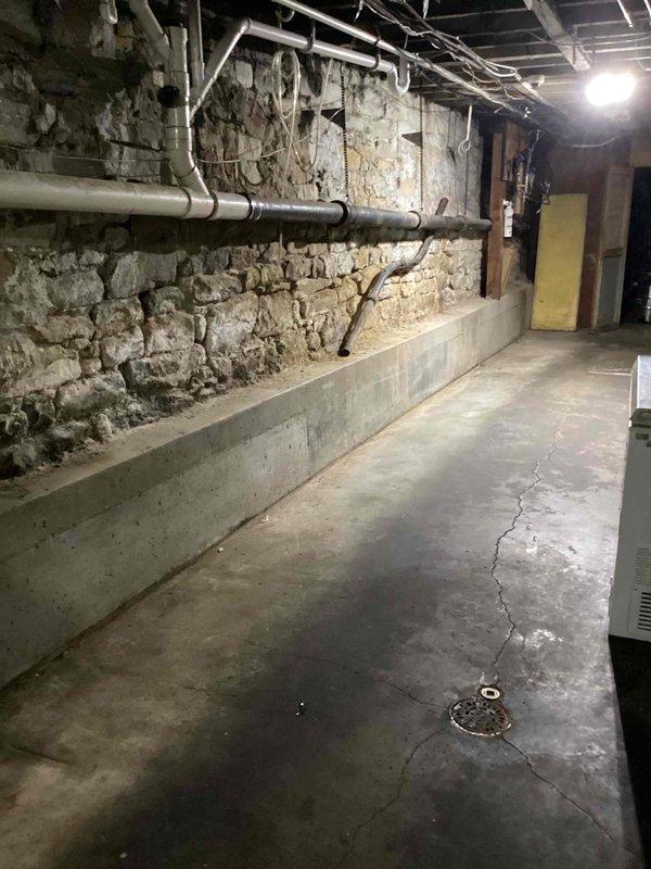 Conducted site assessment of commercial kitchen basement area to evaluate existing plumbing infrastructure and provide pricing for complete kitchen renovation. Documented current conditions including aged stone foundation walls, exposed utility lines, concrete flooring with drainage systems, and horizontal PVC piping mounted along perimeter walls. Photographed basement mechanical room showing deteriorated concrete surfaces, corroded horizontal piping, and existing HVAC equipment placement to determine scope of plumbing modifications required for kitchen remodel project.