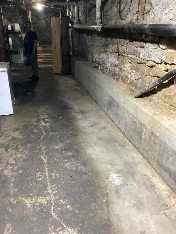 Conducted site assessment of commercial kitchen basement area to evaluate existing plumbing infrastructure and provide pricing for complete kitchen renovation. Documented current conditions including aged stone foundation walls, exposed utility lines, concrete flooring with drainage systems, and horizontal PVC piping mounted along perimeter walls. Photographed basement mechanical room showing deteriorated concrete surfaces, corroded horizontal piping, and existing HVAC equipment placement to determine scope of plumbing modifications required for kitchen remodel project.