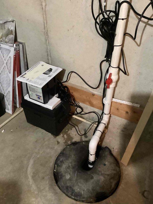 Installed sump pump system with white PVC discharge piping in basement utility area per customer specifications. Configured vertical discharge line from sump pit with proper fittings and cleanout access, secured horizontal pipe runs along ceiling joists using appropriate hangers and support clamps, and completed electrical connections for pump operation.