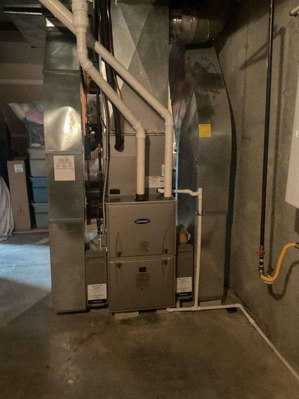 Completed full HVAC system installation in residential basement mechanical room. Installed new Carrier brand furnace unit with proper venting configuration using white PVC intake and exhaust pipes routed vertically through structure. Mounted silver air handler unit on concrete floor with evaporator coil assembly positioned above handler. Connected gray metal supply plenum and ductwork transitions to distribution system. Ran yellow flexible gas line to furnace per code requirements. Installed tall gray metal return air ductwork component for proper air circulation. Secured all manufacturer labels and documentation to equipment faces for reference. Verified all electrical connections and wiring terminations. Tested system startup sequence and confirmed proper operation of all components. System commissioned and left in full operational status providing heating and cooling capacity to residence.