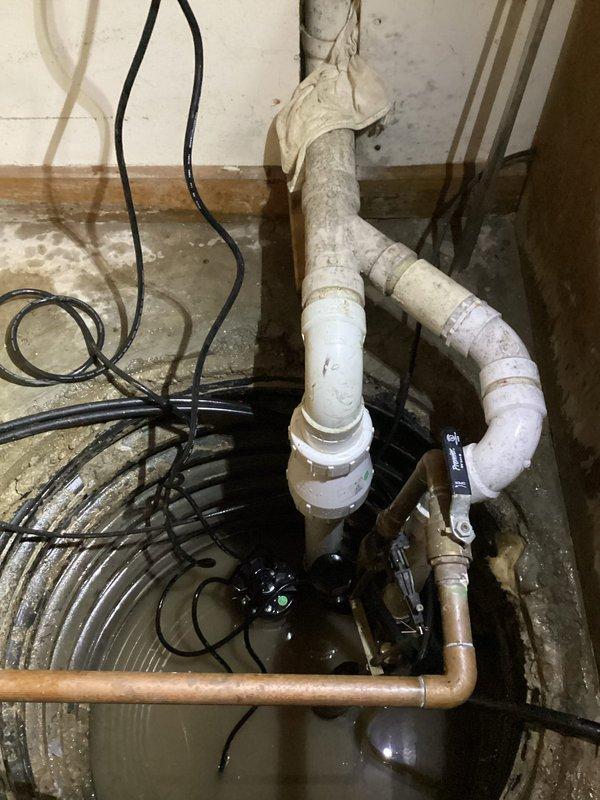 Responded to sump pump malfunction after water company detected excessive water discharge of thousands of gallons and shut off the system. Upon arrival, inspected sump pit containing standing water with submersible pump partially submerged in murky water. Evaluated complete pump assembly including vertical white PVC discharge piping secured to concrete wall, horizontal copper connections, and multiple drainage pipe branches. Assessed electrical wiring and power connections for proper operation. Documented existing installation showing signs of age with discoloration on PVC and copper components, water staining on surrounding concrete floor, and deterioration around pit perimeter. Photographed pump system configuration and standing water condition in ribbed plastic pit liner for service records.