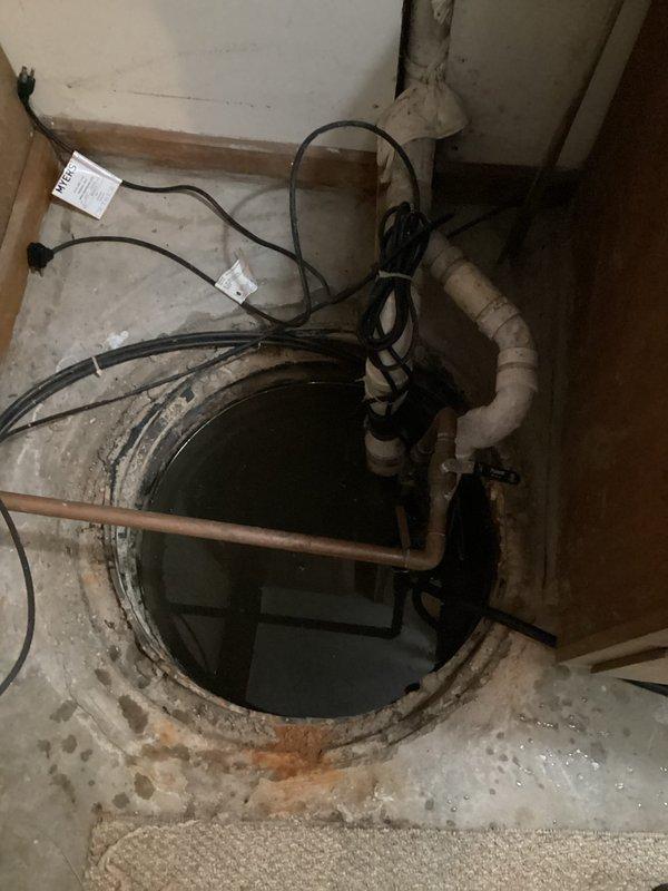 Responded to sump pump malfunction after water company detected excessive water discharge of thousands of gallons and shut off the system. Upon arrival, inspected sump pit containing standing water with submersible pump partially submerged in murky water. Evaluated complete pump assembly including vertical white PVC discharge piping secured to concrete wall, horizontal copper connections, and multiple drainage pipe branches. Assessed electrical wiring and power connections for proper operation. Documented existing installation showing signs of age with discoloration on PVC and copper components, water staining on surrounding concrete floor, and deterioration around pit perimeter. Photographed pump system configuration and standing water condition in ribbed plastic pit liner for service records.