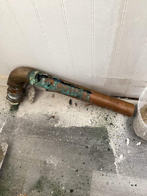 Responded to water leak investigation with vanity already removed by client. Accessed wall cavity to locate source of leak behind three-handle tub/shower valve assembly. Identified corroded copper supply piping at 90-degree elbow joint showing extensive verdigris oxidation and active leak point. Removed deteriorated section of copper pipe and fittings. Installed new copper pipe and elbow joint with proper soldered connections. Pressure tested repair to verify leak elimination. Wall showed significant water damage with rust staining around fixture mounting area, indicating prolonged moisture exposure. Documented pre-existing conditions including corroded plumbing, water-damaged beadboard paneling, and moisture staining on concrete floor. Repair completed successfully with no active leaks detected upon final inspection.