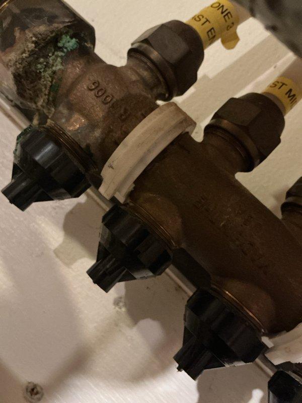 Responded to service call for hydronic boiler system experiencing active water leaks, mineral deposits, and high-pitched whining noise. Upon inspection, found extensive corrosion and oxidation on zone valve bodies and manifold components, with visible turquoise-green patina indicating prolonged moisture exposure. Identified multiple dripping connections throughout the distribution manifold serving the radiant heating zones. Documented condition of aged brass valves showing significant surface deterioration and mineral buildup on copper piping. Water staining observed on floor beneath manifold assembly confirmed active leaking from system components. Photographed corroded valve assemblies and multi-zone manifold system to document extent of deterioration requiring repair or replacement.