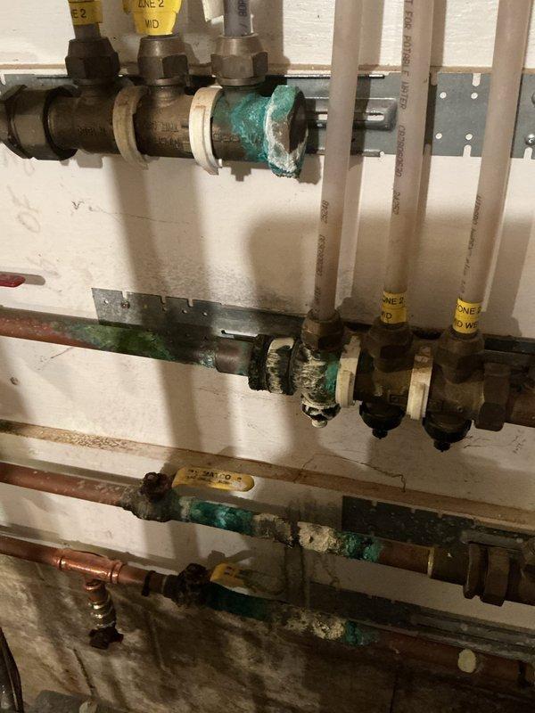 Responded to service call for hydronic boiler system experiencing active water leaks, mineral deposits, and high-pitched whining noise. Upon inspection, found extensive corrosion and oxidation on zone valve bodies and manifold components, with visible turquoise-green patina indicating prolonged moisture exposure. Identified multiple dripping connections throughout the distribution manifold serving the radiant heating zones. Documented condition of aged brass valves showing significant surface deterioration and mineral buildup on copper piping. Water staining observed on floor beneath manifold assembly confirmed active leaking from system components. Photographed corroded valve assemblies and multi-zone manifold system to document extent of deterioration requiring repair or replacement.