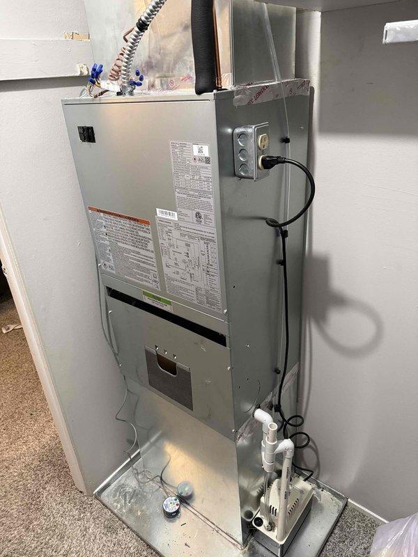 Completed installation of new HVAC air handler unit with evaporator coil in residential utility closet. Positioned and secured gray metal cabinet-style air handler against wall, ensuring proper clearances and structural support. Connected flexible metal ductwork from unit to existing plenum system, verifying proper sealing and airflow configuration. Installed electrical disconnect box on adjacent wall and completed all power wiring connections per manufacturer specifications and local electrical codes. Mounted and plumbed white condensate pump assembly at base of unit with appropriate drainage tubing to handle moisture removal during cooling operation. Verified all manufacturer labels, warning decals, and specification plates are visible and properly affixed to unit. Connected black flexible supply duct from top of air handler to ceiling distribution system, ensuring secure attachment and proper insulation interface. Tested all electrical connections for proper voltage and amperage. Co
