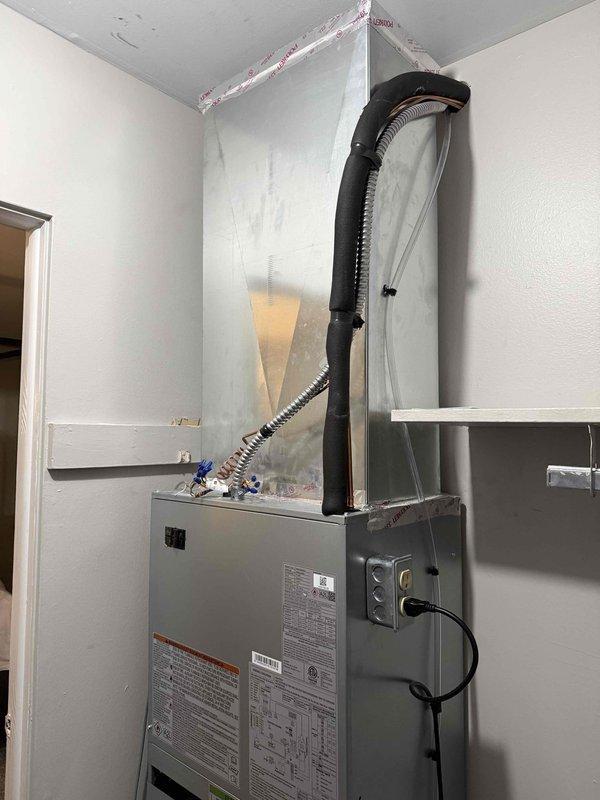 Completed installation of new HVAC air handler unit with evaporator coil in residential utility closet. Positioned and secured gray metal cabinet-style air handler against wall, ensuring proper clearances and structural support. Connected flexible metal ductwork from unit to existing plenum system, verifying proper sealing and airflow configuration. Installed electrical disconnect box on adjacent wall and completed all power wiring connections per manufacturer specifications and local electrical codes. Mounted and plumbed white condensate pump assembly at base of unit with appropriate drainage tubing to handle moisture removal during cooling operation. Verified all manufacturer labels, warning decals, and specification plates are visible and properly affixed to unit. Connected black flexible supply duct from top of air handler to ceiling distribution system, ensuring secure attachment and proper insulation interface. Tested all electrical connections for proper voltage and amperage. Co