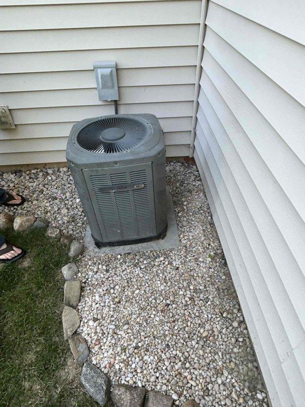 Completed HVAC system replacement under tech lead Joel's supervision. Installed new Trane furnace unit in basement utility area with associated ductwork, including metallic silver ducting with curved elbow joints and proper hose clamp connections. Replaced outdoor air conditioning unit positioned in narrow alcove between house sections, mounted on concrete pad with proper electrical connections. System includes white air handling components, copper refrigerant lines, electrical conduit, and wall-mounted controls, all installed to manufacturer specifications and code requirements.