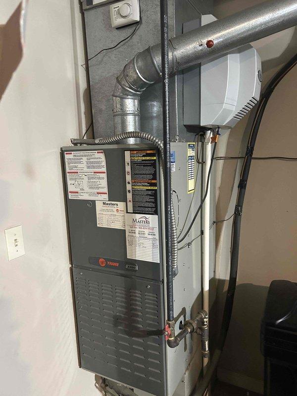 Completed HVAC system replacement under tech lead Joel's supervision. Installed new Trane furnace unit in basement utility area with associated ductwork, including metallic silver ducting with curved elbow joints and proper hose clamp connections. Replaced outdoor air conditioning unit positioned in narrow alcove between house sections, mounted on concrete pad with proper electrical connections. System includes white air handling components, copper refrigerant lines, electrical conduit, and wall-mounted controls, all installed to manufacturer specifications and code requirements.