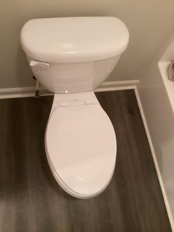 Completed installation of customer-supplied two-piece white porcelain toilet in residential bathroom. Removed existing fixture, installed new toilet with proper wax ring seal, secured unit to floor flange with closet bolts, and connected water supply line. Verified proper flush operation, checked for leaks at all connection points, and confirmed stable mounting. Toilet installed on dark vinyl plank flooring between bathtub and wall with appropriate clearances maintained.