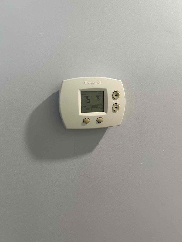 Conducted HVAC system evaluation for equipment replacement estimate. Documented existing Honeywell digital thermostat displaying 75 degrees and measured ductwork dimensions at approximately 22-24 inches for proper sizing calculations. Photographed current equipment configuration including cylindrical duct components and cabinet housing to assess replacement requirements and ensure accurate specifications for new system installation.