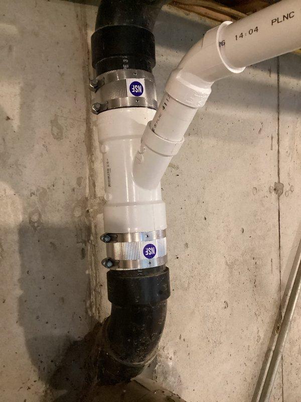 Installed new sump pump system with complete discharge line assembly in unfinished basement utility area. The installation included white PVC piping with professional-grade metallic coupling bands secured with hose clamps, configured with multiple 90-degree elbows to route discharge line along foundation wall and up through ceiling joists. Connected pump to black flexible discharge piping, ensuring proper flow direction and secure watertight connections throughout the system.