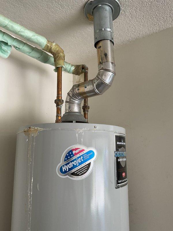 Responded to service call for leaking tanked gas water heater in garage with visible water accumulation around base and in drain pan. Upon inspection, identified active leak at bottom of unit with moisture staining on concrete floor and corrosion present on metal support frame base. Documented existing Hydrojet brand water heater installation including plumbing connections, copper piping with foam insulation, PVC venting system, and drain pan configuration. Photographed leak source, water damage extent, and overall installation condition for assessment and customer records.
