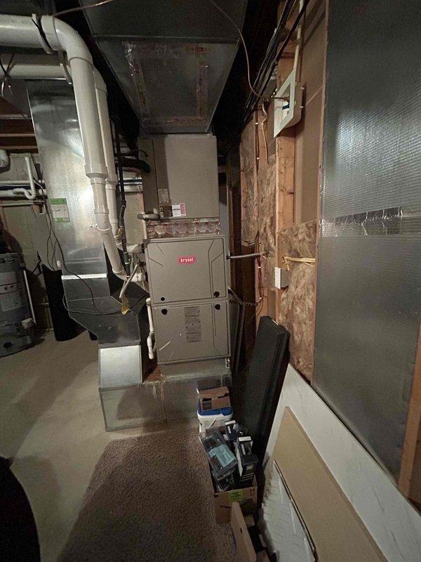 Performed comprehensive site evaluation for complete HVAC system replacement at residential property experiencing no cooling condition. Assessed existing outdoor condensing unit positioned on grade-level concrete pad against vinyl siding, noting weathered equipment with accumulated debris at base and standard electrical service connection via gray conduit and junction box. Evaluated indoor Bryant furnace system located in basement mechanical room with existing ductwork, PVC venting, and copper refrigerant lines. Documented current equipment specifications, electrical service requirements, and installation access points. Photographed existing equipment conditions and mechanical room layout to prepare detailed replacement estimate for new cooling and heating system installation.