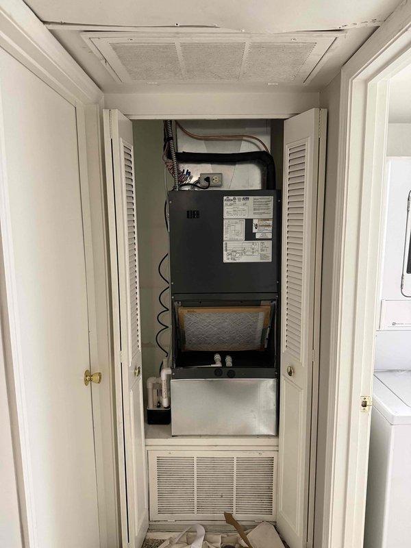 Completed installation of customer-supplied air conditioning system and air handler unit in end closet location. The new black air handler was mounted vertically in the utility closet with proper ductwork connections, condensate pan, and filtration system installed. Exterior condenser units were positioned on gravel pad against stone wall with appropriate clearances and electrical disconnects mounted. Removed and transported old HVAC equipment to maintenance office for disposal.
