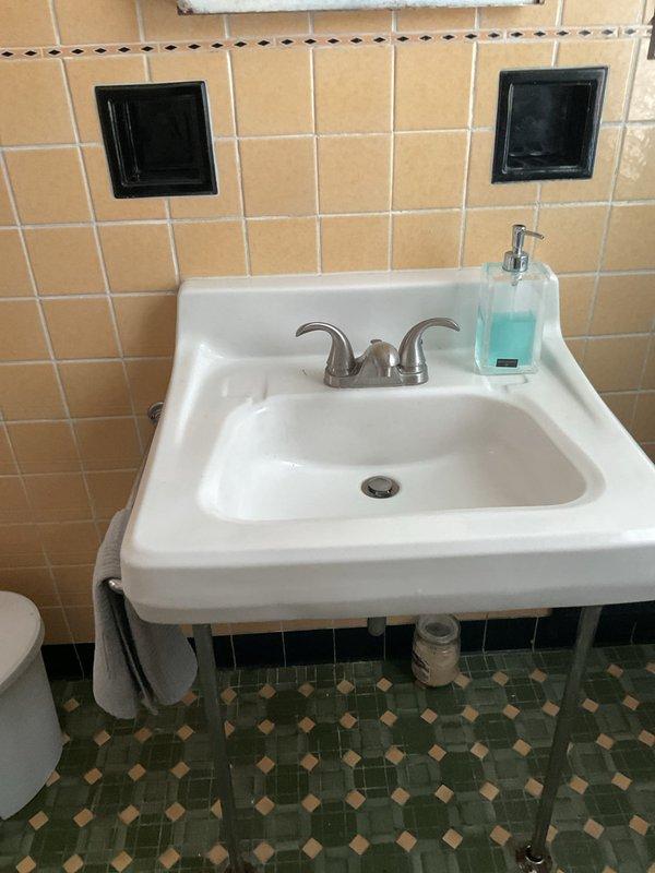 Conducted on-site assessment for bathroom fixture replacement in downstairs bathroom. Evaluated existing white pedestal sink with chrome dual-handle faucet mounted against beige tiled wall with decorative diamond border and recessed soap niches. Documented current installation including exposed P-trap plumbing and green tile flooring with diamond accent pattern. Reviewed replacement vanity unit specifications featuring white rectangular integrated sink basin with three pre-drilled faucet holes, white countertop, and light wood cabinet base with drawer and open shelving storage. Provided estimate for complete fixture removal and new vanity installation including necessary plumbing connections and drain assembly.