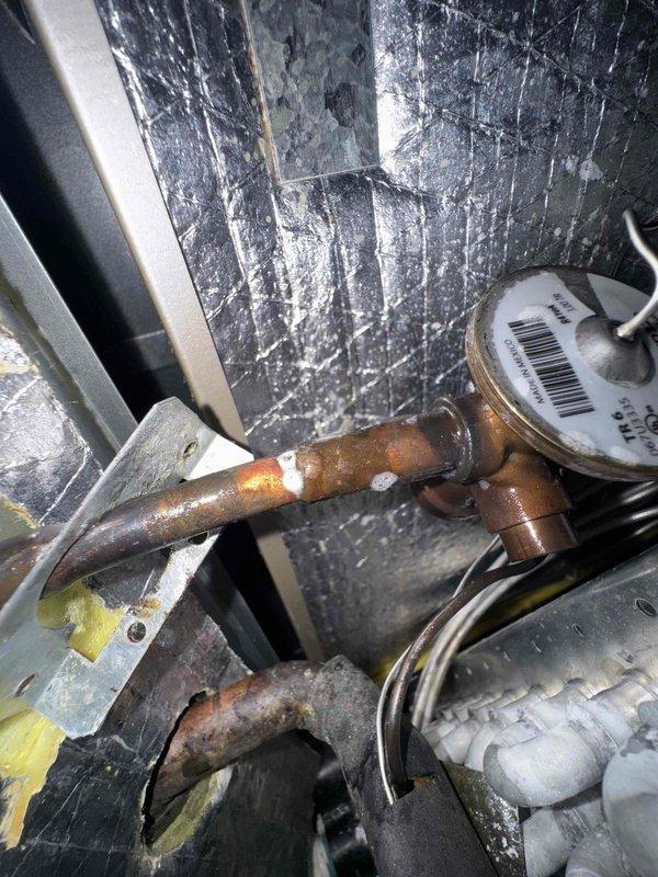 Responded to callback for no cooling condition with outdoor unit short cycling and water accumulation around furnace following recent TXV replacement. Upon inspection, discovered severe corrosion and rust on refrigerant piping connections, particularly on vertical pipe sections near copper fittings and T-joints within air handler cabinet. Corrosion exhibited active deterioration with heavy oxidation present on multiple copper line sections. Documented extensive pipe degradation and compromised connections that likely contributed to system malfunction and condensate issues. Photographed corroded refrigerant lines and deteriorated fittings for reference and potential warranty consideration related to previous repair work.