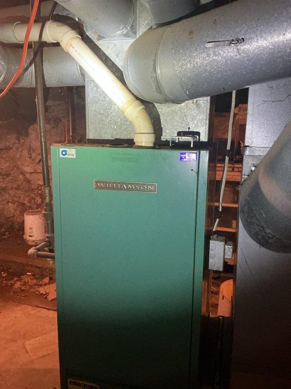 Conducted on-site assessment of 32-year-old Williamson propane gas furnace system located in basement utility area. Unit showing significant age-related wear and approaching end of serviceable life. Documented existing equipment configuration including green cabinet heating unit, PVC venting system, metallic ductwork with visible condensation, and basement installation environment. Photographed current system setup, floor register placement, gas piping with shut-off valve, and surrounding mechanical space conditions. Provided comprehensive replacement estimate for new propane furnace system to address aging equipment concerns and ensure continued reliable heating operation.