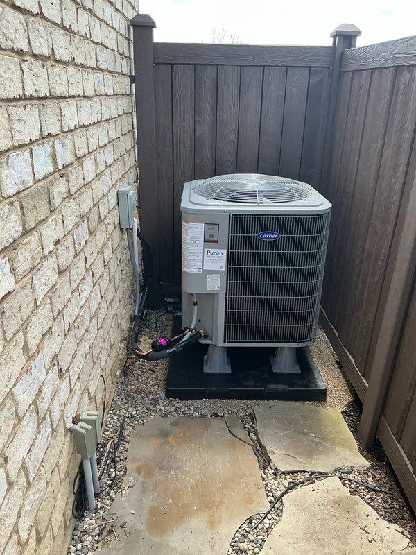 Completed full HVAC system installation including Carrier 27SCA524A003 outdoor condensing unit and FJ5ANXB24L00 indoor air handler. The outdoor unit was positioned on a composite pad in the side yard area between the residence and privacy fence, with proper clearances maintained and electrical conduit securely mounted to exterior wall. The indoor air handler was installed in the basement utility area within wooden framing structure, with all manufacturer labels and specifications clearly visible and accessible for future service requirements.