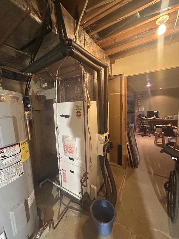 Conducted on-site assessment of existing HVAC system in basement utility area to provide replacement estimate. Evaluated current white furnace/air handler unit and associated ductwork configuration. Documented equipment condition, noting existing installation includes gray water heater adjacent to HVAC unit with metal piping and ductwork running along exposed ceiling joists. Observed evidence of water damage on concrete floor and active moisture concerns with bucket positioned for collection, which may impact equipment longevity and replacement planning. Photographed current system layout, equipment specifications, and utility area conditions to prepare comprehensive replacement quote for new air conditioning system.