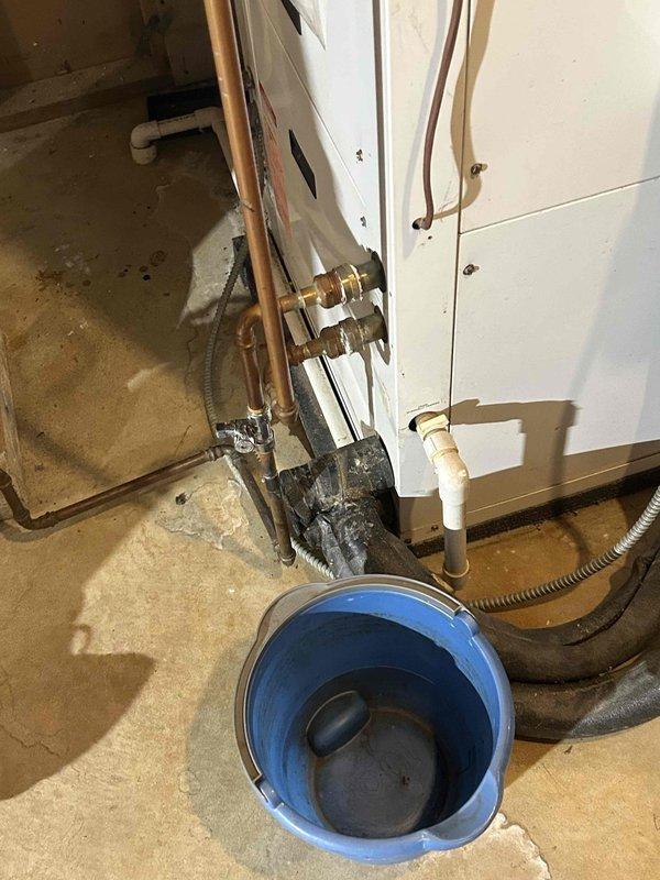 Conducted on-site assessment of existing HVAC system in basement utility area to provide replacement estimate. Evaluated current white furnace/air handler unit and associated ductwork configuration. Documented equipment condition, noting existing installation includes gray water heater adjacent to HVAC unit with metal piping and ductwork running along exposed ceiling joists. Observed evidence of water damage on concrete floor and active moisture concerns with bucket positioned for collection, which may impact equipment longevity and replacement planning. Photographed current system layout, equipment specifications, and utility area conditions to prepare comprehensive replacement quote for new air conditioning system.