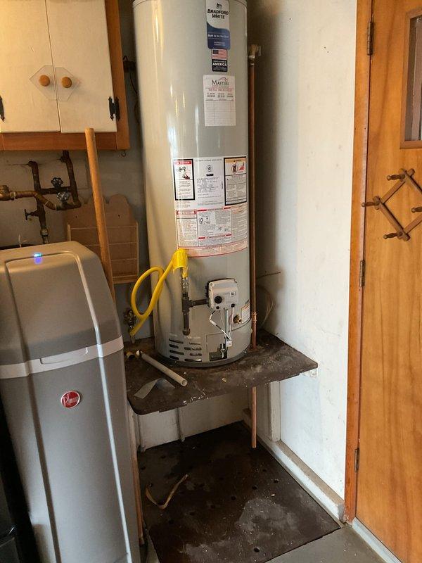 Successfully installed a new 40-gallon natural gas tank water heater in garage utility area. Unit was positioned on existing metal stand platform in tight-fit location adjacent to water softener system. Connected gas supply line using flexible yellow gas connector and completed all necessary copper water line connections. Verified proper clearances and measurements for equipment placement. Installation required additional assistance due to confined space and need to position unit securely on elevated stand.