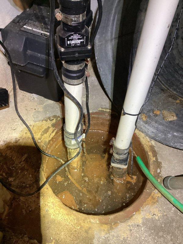 Responded to service call for non-operational sump pump system. Upon arrival, inspected sump pit installation containing standing water with brownish discoloration, indicating potential drainage issues. Evaluated complete system including black cylindrical pump motor mounted vertically in circular basin, white PVC discharge piping with U-shaped configuration, darker secondary pipe, and electrical control box with power connections. Assessed pump mounting hardware including metal hose clamps and zip tie securing mechanisms. Examined electrical components, wiring connections, and control unit mounted to wall structure. Inspected discharge piping configuration and verified proper installation of all plumbing connections. Noted evidence of water staining around pit opening and corrugated drainage components. Documented system condition through photographic records showing pump assembly, piping arrangement, electrical controls, and surrounding basement environment with visible concrete wall