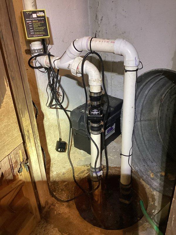 Responded to service call for non-operational sump pump system. Upon arrival, inspected sump pit installation containing standing water with brownish discoloration, indicating potential drainage issues. Evaluated complete system including black cylindrical pump motor mounted vertically in circular basin, white PVC discharge piping with U-shaped configuration, darker secondary pipe, and electrical control box with power connections. Assessed pump mounting hardware including metal hose clamps and zip tie securing mechanisms. Examined electrical components, wiring connections, and control unit mounted to wall structure. Inspected discharge piping configuration and verified proper installation of all plumbing connections. Noted evidence of water staining around pit opening and corrugated drainage components. Documented system condition through photographic records showing pump assembly, piping arrangement, electrical controls, and surrounding basement environment with visible concrete wall