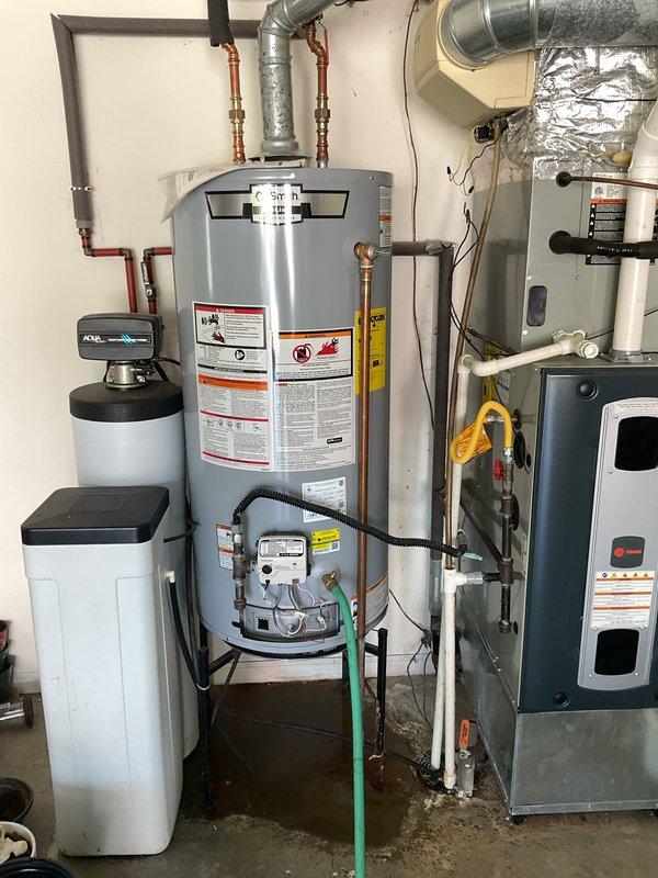 Responded to service call for leaking tanked gas water heater located in garage utility area. Upon arrival, performed visual inspection and identified active water leak originating from base of unit with visible moisture damage and water staining on concrete floor beneath heater. Documented existing installation including water heater, water softener, and HVAC components with connected copper piping systems. Assessed condition of tank, gas valve assembly, temperature and pressure relief valve, and all associated plumbing connections. Photographed leak evidence and overall equipment setup for service documentation and customer records.