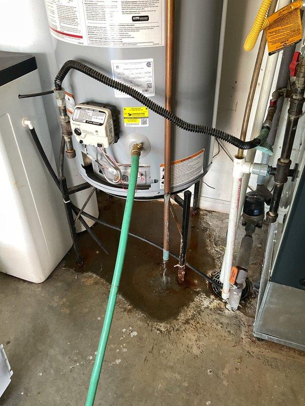 Responded to service call for leaking tanked gas water heater located in garage utility area. Upon arrival, performed visual inspection and identified active water leak originating from base of unit with visible moisture damage and water staining on concrete floor beneath heater. Documented existing installation including water heater, water softener, and HVAC components with connected copper piping systems. Assessed condition of tank, gas valve assembly, temperature and pressure relief valve, and all associated plumbing connections. Photographed leak evidence and overall equipment setup for service documentation and customer records.