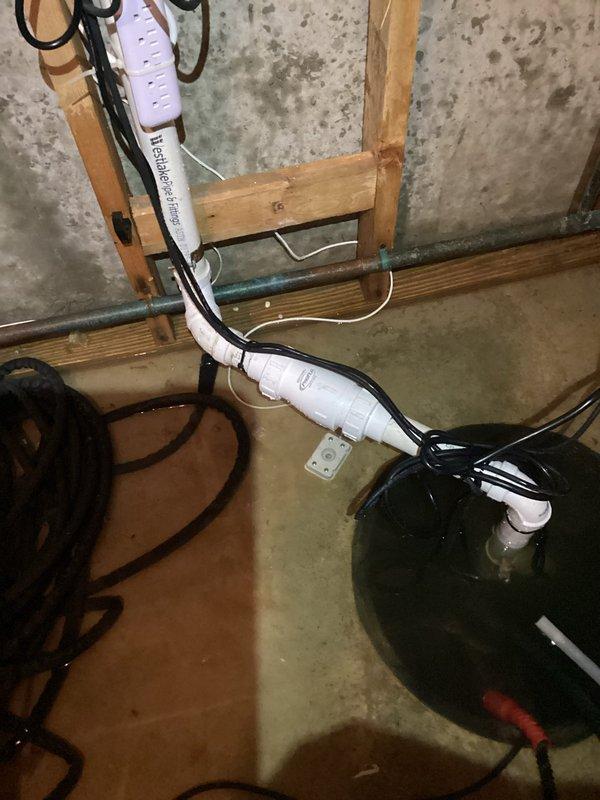 Responded to service call for sump pump system failure causing water backup into residence. Upon inspection, found dual sump pump installation in basement with inadequate pumping capacity and operational issues preventing proper water removal. Assessed discharge system including white PVC piping and electrical connections with power strip configuration. Identified compromised pump performance leading to water accumulation on basement floor, evidenced by visible staining and moisture marks on concrete surfaces. Verified discharge line routing to exterior property where water exits into drainage area near lawn perimeter. Addressed pump functionality concerns to restore proper water evacuation and prevent further basement flooding.