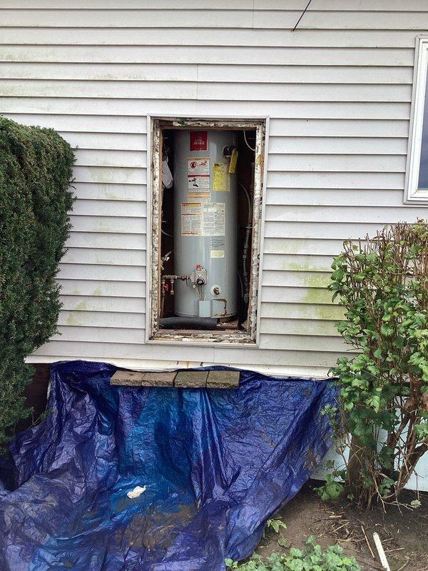 Provided estimate for gas water heater replacement in mobile home. Unit is currently accessed through exterior wall opening with visible deterioration to wooden frame requiring attention during installation. Assessed existing installation configuration including gas supply lines, venting requirements, and exterior access panel specifications unique to mobile home applications.