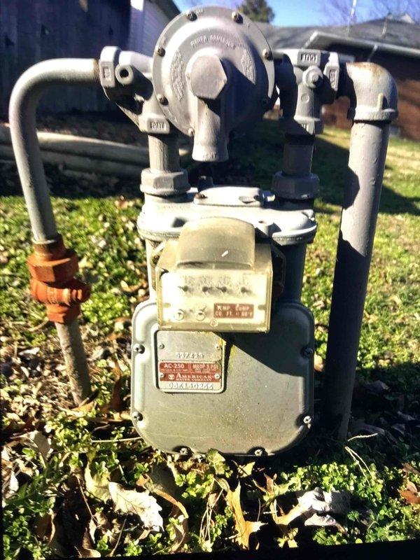 Conducted site assessment and documented existing gas meter installation for replacement estimate. Current system shows an aging American-branded gas meter with visible weathering, connected to both gray metal pipes and copper fittings. Photographed complete setup including pressure regulator assembly and valve components to develop comprehensive upgrade proposal.