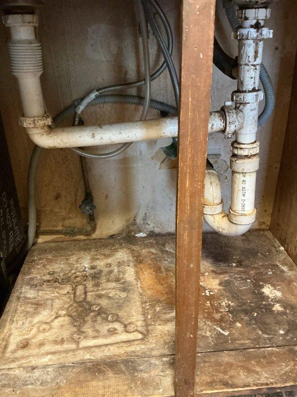 Cleared severely clogged kitchen sink drain by accessing and cleaning the PVC drain assembly under the sink cabinet, which showed significant water damage from previous leakage. Inspected exposed wall section with horizontal plumbing runs to verify proper drainage flow and ensure no additional blockages remained in the system.