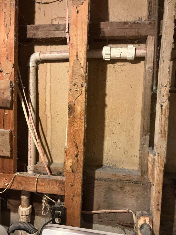 Cleared severely clogged kitchen sink drain by accessing and cleaning the PVC drain assembly under the sink cabinet, which showed significant water damage from previous leakage. Inspected exposed wall section with horizontal plumbing runs to verify proper drainage flow and ensure no additional blockages remained in the system.