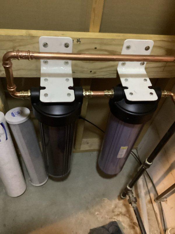 Successfully installed a comprehensive whole-house water filtration system consisting of dual black cylindrical filter housings with white bracket supports securely mounted to the wall structure. Connected the system to existing copper plumbing infrastructure using brass fittings and valves, ensuring proper integration with the home's water supply network including PEX connections where appropriate.