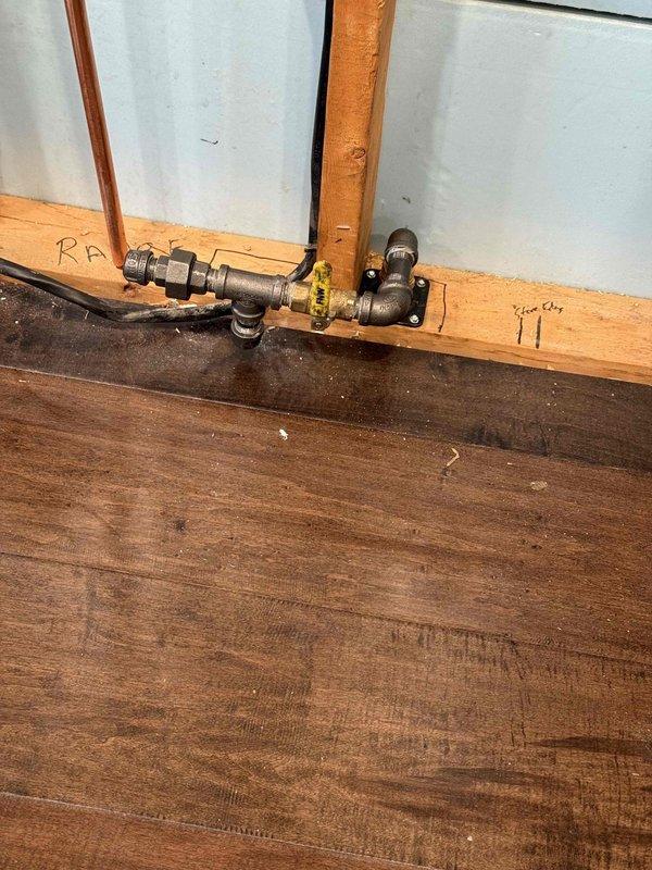 Completed installation of gas lines per sales agreement. Work included mounting a plumbing assembly with T-junction and shut-off valve on wooden baseboard, and installing exterior metal pipe configuration with red valve handle at the brick wall perimeter. All connections were properly secured and tested for functionality.