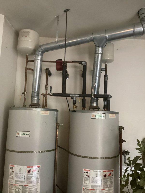 # Dual Water Heater System Installation

Completed professional installation of dual State brand residential water heaters in dedicated mechanical room. Configured units in parallel arrangement to provide increased hot water capacity and redundancy for the residence. 

Installed comprehensive copper and black iron piping manifold system connecting both water heaters with proper isolation valves for independent service capability. Mounted multiple expansion tanks along horizontal pipe runs to accommodate thermal expansion and maintain system pressure stability. Integrated red-handled circulator pump in central piping network to facilitate efficient water distribution between units.

Routed silver galvanized exhaust ductwork from each unit with proper elbow configurations for code-compliant venting. Connected all water supply lines using brass fittings and shut-off valves at strategic locations throughout the system. Installed ceiling-mounted support brackets and hangers to secure all pi