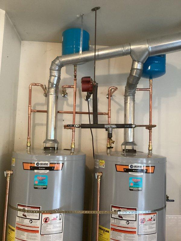 # Dual Water Heater System Installation

Completed professional installation of dual State brand residential water heaters in dedicated mechanical room. Configured units in parallel arrangement to provide increased hot water capacity and redundancy for the residence. 

Installed comprehensive copper and black iron piping manifold system connecting both water heaters with proper isolation valves for independent service capability. Mounted multiple expansion tanks along horizontal pipe runs to accommodate thermal expansion and maintain system pressure stability. Integrated red-handled circulator pump in central piping network to facilitate efficient water distribution between units.

Routed silver galvanized exhaust ductwork from each unit with proper elbow configurations for code-compliant venting. Connected all water supply lines using brass fittings and shut-off valves at strategic locations throughout the system. Installed ceiling-mounted support brackets and hangers to secure all pi