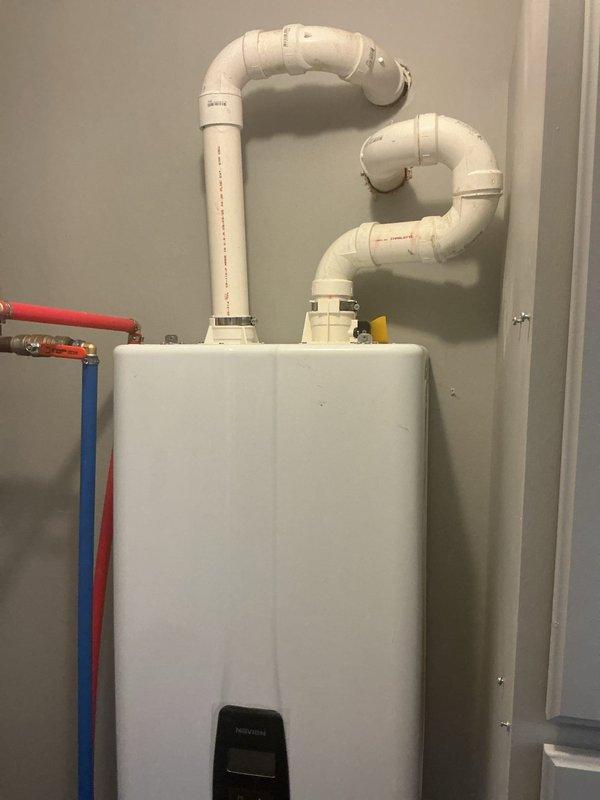 Completed professional installation and inspection of wall-mounted tankless water heater system in residential utility space. Installation includes properly configured white PVC venting with S-shaped exhaust loops extending to ceiling, color-coded PEX supply lines (red for hot water, blue for cold water), copper piping connections, yellow flexible gas supply line, and blue ball valve shut-off controls. Unit displays ENERGY STAR certification and features digital control panel for temperature management. All plumbing connections secured to wall per manufacturer specifications, with proper valve placement for system maintenance and emergency shut-off capability. Venting configuration meets code requirements for gas-fired tankless operation, ensuring safe exhaust of combustion gases. System tested and verified operational.
