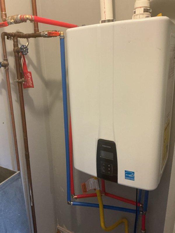 Completed professional installation and inspection of wall-mounted tankless water heater system in residential utility space. Installation includes properly configured white PVC venting with S-shaped exhaust loops extending to ceiling, color-coded PEX supply lines (red for hot water, blue for cold water), copper piping connections, yellow flexible gas supply line, and blue ball valve shut-off controls. Unit displays ENERGY STAR certification and features digital control panel for temperature management. All plumbing connections secured to wall per manufacturer specifications, with proper valve placement for system maintenance and emergency shut-off capability. Venting configuration meets code requirements for gas-fired tankless operation, ensuring safe exhaust of combustion gases. System tested and verified operational.