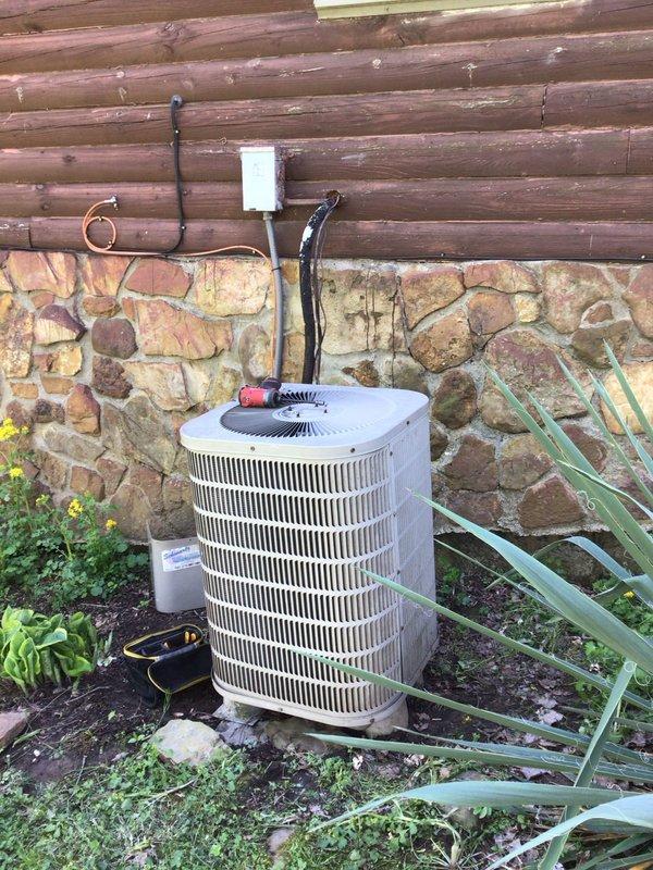 Responded to no-cooling complaint on 9+ year old residential air conditioning system. Customer reported unit was running hot with abnormal humming noise before shutting system down. Upon arrival, performed diagnostic evaluation of outdoor condensing unit located at exterior log cabin wall. Inspected cylindrical-style condenser with vertical grille slats, refrigerant lines, and electrical disconnect. Evaluated compressor operation, capacitor function, and electrical components to identify source of humming noise and overheating condition. Checked refrigerant pressures, amperage draws, and contactor operation. Assessed airflow through condenser coil and verified proper fan motor operation. System was found to be inoperative with no breakers tripped at electrical panel. Documented equipment condition with photos showing unit placement, surrounding landscape vegetation, and mounting configuration against stone foundation. Completed thorough system diagnostics to determine root cause of coo