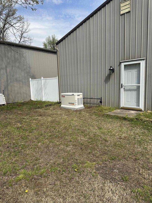 # Electrical Install - Generator

Successfully completed installation of a 26kW Generac standby generator system for residential application. Unit was mounted on a concrete pad adjacent to metal-sided building structure with proper clearances maintained per manufacturer specifications and local code requirements. Installed transfer switch and completed all necessary electrical connections from main service panel to generator unit. Black conduit runs were properly secured along building exterior to house electrical wiring between components. Generator cabinet positioned for optimal ventilation and serviceability access. System tested and verified operational, ready for automatic backup power operation during utility outages.