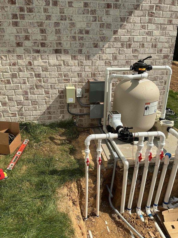 Completed electrical installation for new pool equipment system. Installed conduit and wiring from green electrical control box mounted on exterior brick wall to pool filtration equipment, including connections for sand filter tank with black valve assembly and manifold piping system with multiple ball valves. All electrical connections made to code specifications to support pool pump and filtration system operation.