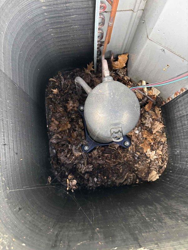 Completed first annual maintenance visit on a residential HVAC system over 20 years old. Performed comprehensive inspection of outdoor condenser unit, which is protected by a weathered wire cage enclosure with visible corrosion on upper sections. Discovered significant accumulation of decomposed organic debris, primarily leaves and vegetation, within the utility access pit surrounding the sump pump and drainage system at the bottom of the equipment well. Noted standing water present at base of pit. Inspected electrical disconnect box, conduit routing, and refrigerant lines at condenser unit. Documented equipment condition through photographs for maintenance records. System operational at time of service.
