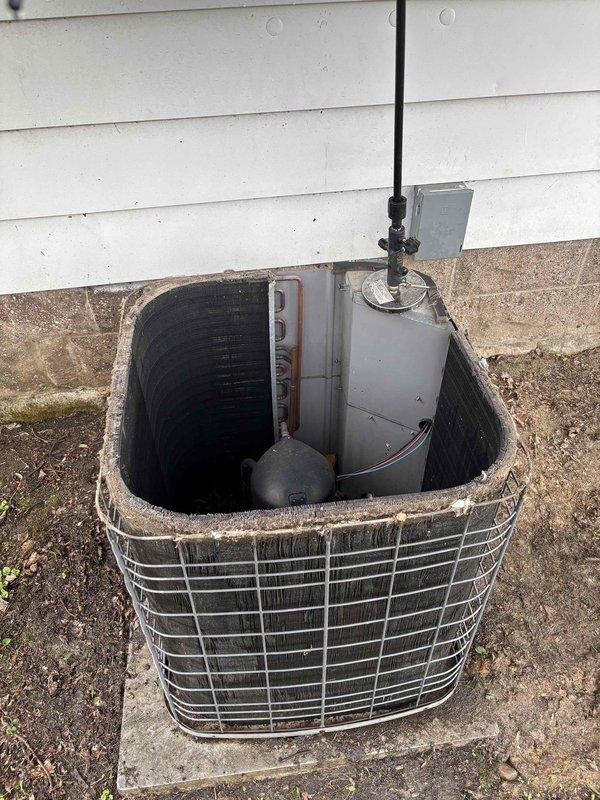 Completed first annual maintenance visit on a residential HVAC system over 20 years old. Performed comprehensive inspection of outdoor condenser unit, which is protected by a weathered wire cage enclosure with visible corrosion on upper sections. Discovered significant accumulation of decomposed organic debris, primarily leaves and vegetation, within the utility access pit surrounding the sump pump and drainage system at the bottom of the equipment well. Noted standing water present at base of pit. Inspected electrical disconnect box, conduit routing, and refrigerant lines at condenser unit. Documented equipment condition through photographs for maintenance records. System operational at time of service.
