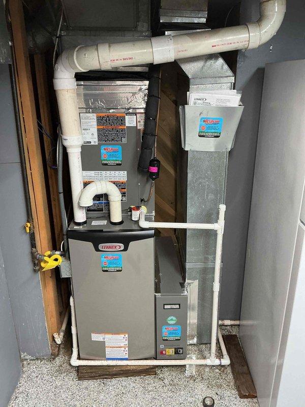 # HVAC Install - Complete System

Successfully completed full HVAC system installation featuring a Lennox furnace unit. The installation was performed in a utility area with proper clearances and professional workmanship throughout.

The furnace was mounted on a white platform base with adequate structural support. Galvanized sheet metal ductwork was installed above the unit to ensure proper airflow distribution. White PVC condensate drain piping was configured in a U-shaped trap on the left side of the furnace to facilitate proper drainage and prevent airflow issues.

Internal components were properly installed and verified, including the blue flame igniter assembly mounted on refractory board with appropriate electrical connections. Gas valve and pressure switch components were secured with proper fittings and connections. All electrical wiring was routed correctly throughout the system, including red, purple, and white conductor wires to burner assembly, blower motor, and control co