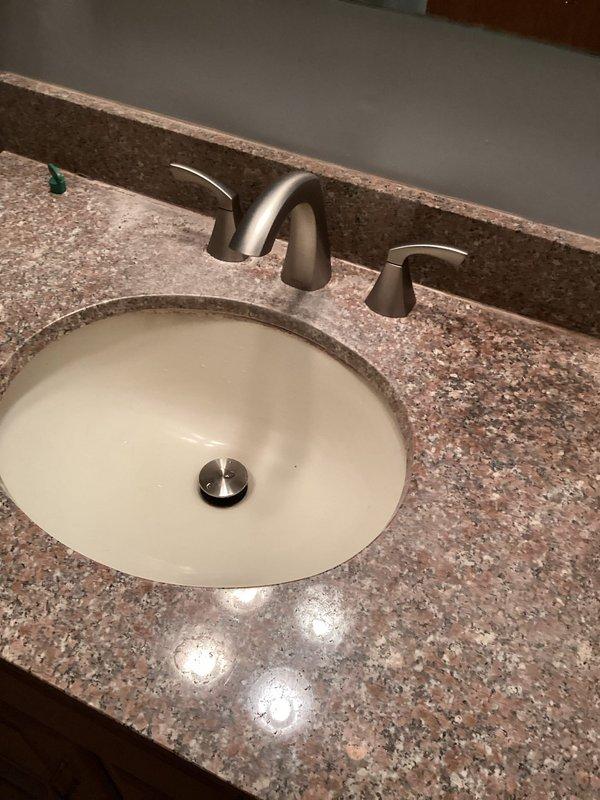 Completed general plumbing installation in residential bathroom, including installation of a two-piece white porcelain toilet with chrome flexible water supply line and wall-mounted shut-off valve in corner placement. Additionally installed undermount white oval sink in granite countertop with three-piece brushed nickel faucet set featuring central spout and dual lever handles, along with polished metal drain assembly.