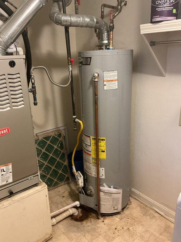 Completed installation of residential water heater in utility room with proper plumbing connections including copper and galvanized piping, pressure relief valve, shut-off valves with red handles, and flexible yellow gas line. All connections were secured with appropriate brass fittings and compression fittings, and the unit was installed adjacent to existing HVAC equipment with adequate clearance for service access and ventilation requirements.