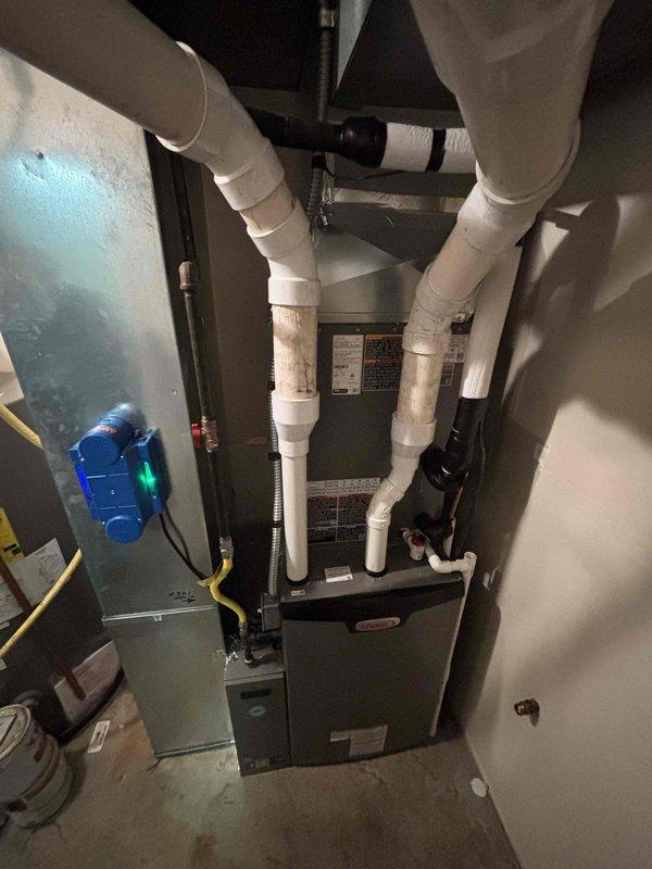 # HVAC Install - Complete System

Successfully completed a full residential HVAC system installation including both indoor and outdoor components. The interior installation features a new furnace unit positioned in the utility area with properly configured white insulated ductwork extending from the unit, secured with metal clamps and routed efficiently across the ceiling space. Installed gray metal plenum with blue gauge monitoring device and yellow flexible coupling connector. All gas and electrical connections completed with black conduit running vertically for proper service access.

The exterior installation includes a new air conditioning condenser unit positioned on a composite pad in the side yard, surrounded by white gravel for drainage and equipment protection. Mounted unit features vertical louvered panels and top-mounted fan assembly. Completed refrigerant line installation with white PVC piping running vertically along the brick exterior wall for proper condensate drainage